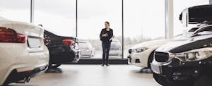 A woman stands in between rows of new cars in an auto dealership showroom.