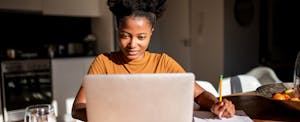 A seated woman looks thoughtfully at her laptop while taking notes.